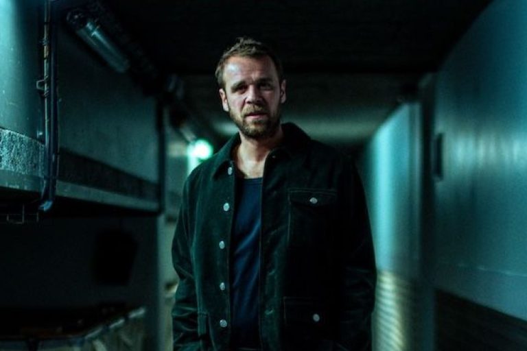 Photo représentant l'artiste Cyril Mokaiesh, debout dans un couloir sombre, regard percant vers l'objectif.