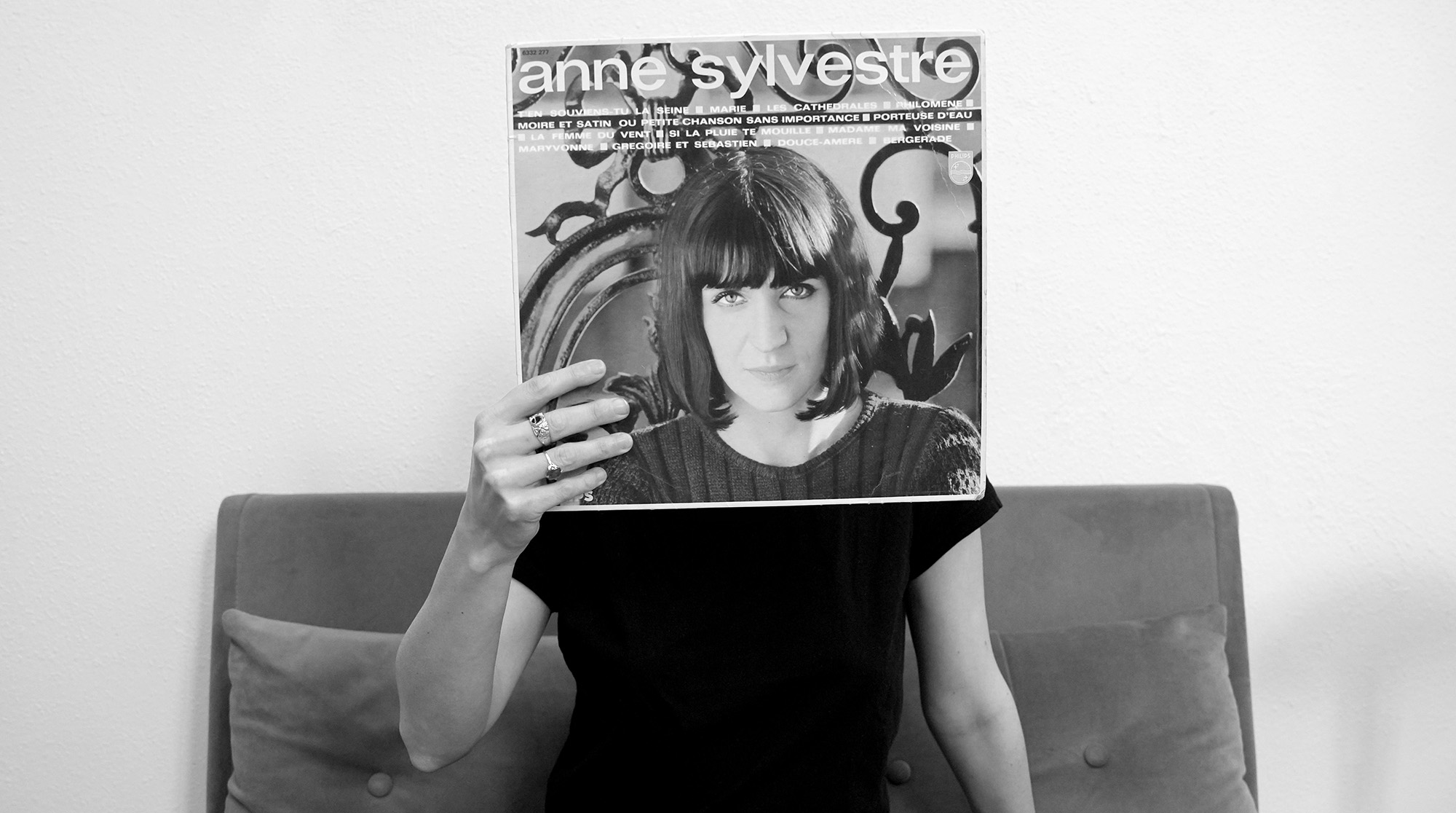 Photo en noir et blanc représentant une jeune femme assise avec une pochette de disque d'Anne Sylvestre devant le visage