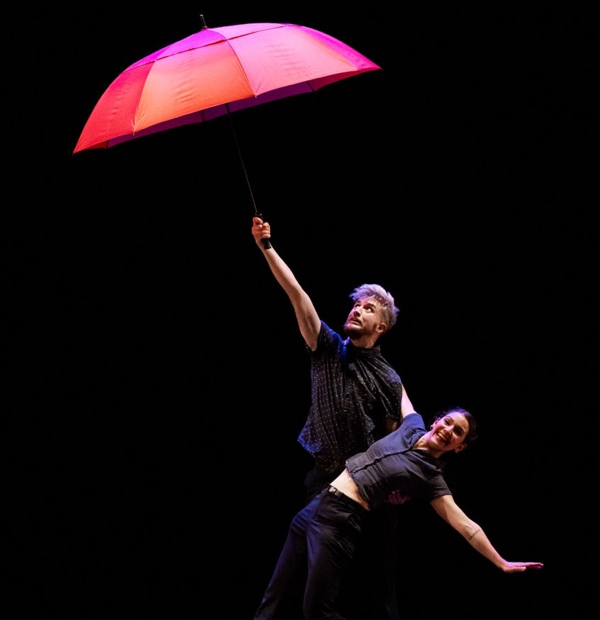 Photo représentant une femme et un homme en train de danser avec un parapluie rouge ouvert.