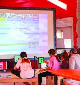 Photo représentant des personnages installées devant un écran géant avec des tablettes, en train de parcourir des photos des Jeux olympiques