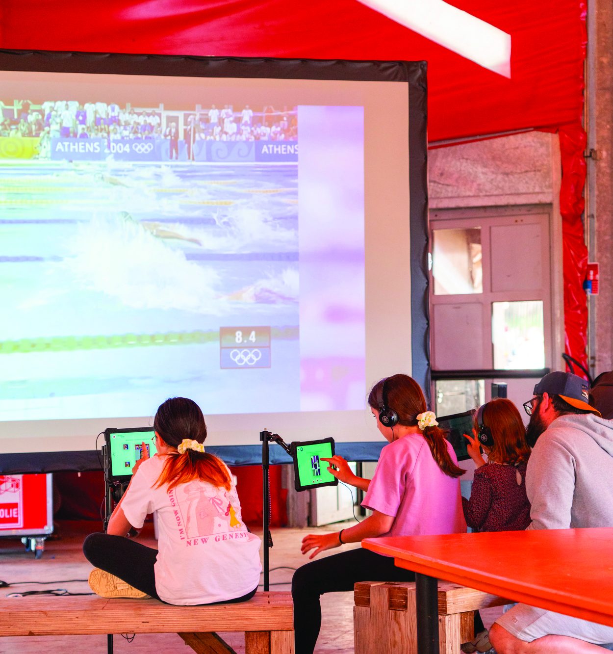 Photo représentant des personnages installées devant un écran géant avec des tablettes, en train de parcourir des photos des Jeux olympiques