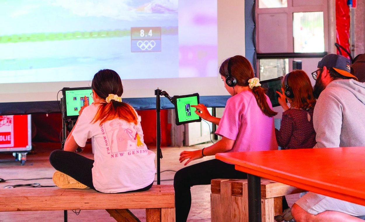 Photo représentant des personnages installées devant un écran géant avec des tablettes, en train de parcourir des photos des Jeux olympiques