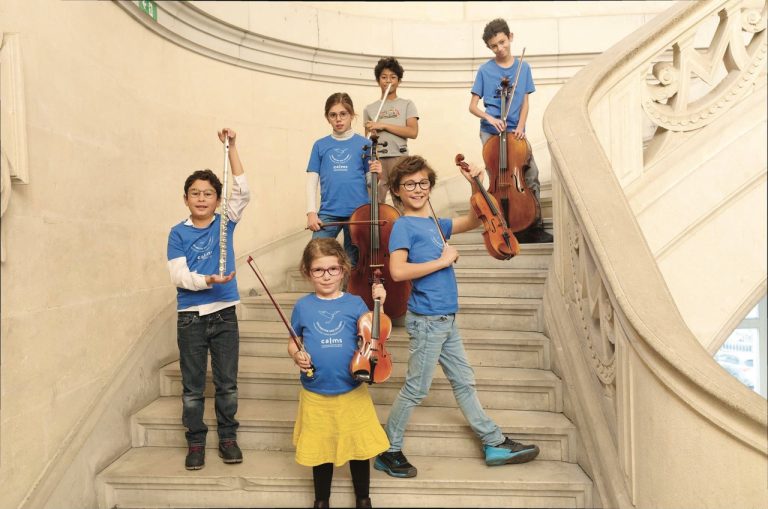 Photo représentant des enfants dans un grand escalier. Ils brandissent fièrement leurs instruments de musique, violon, alto, violoncelle.