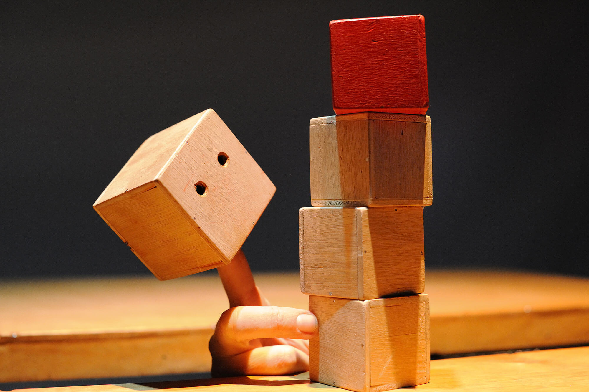 Photo représentant une scène de théâtre d'objets. Un cube en bois orné de deux yeux qui observent des cubes empilés.
