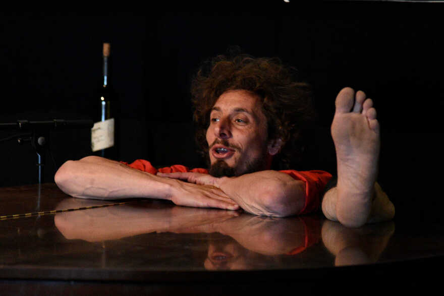 Photo représentant un homme assis, acoudé à une table un pied sur la table et une bouteille à sa gauche.