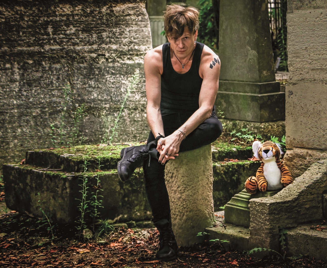 Photo représentant un homme assis sur une pied, jambes croisées. Il porte un pantalon noir et un débrdeur noir.