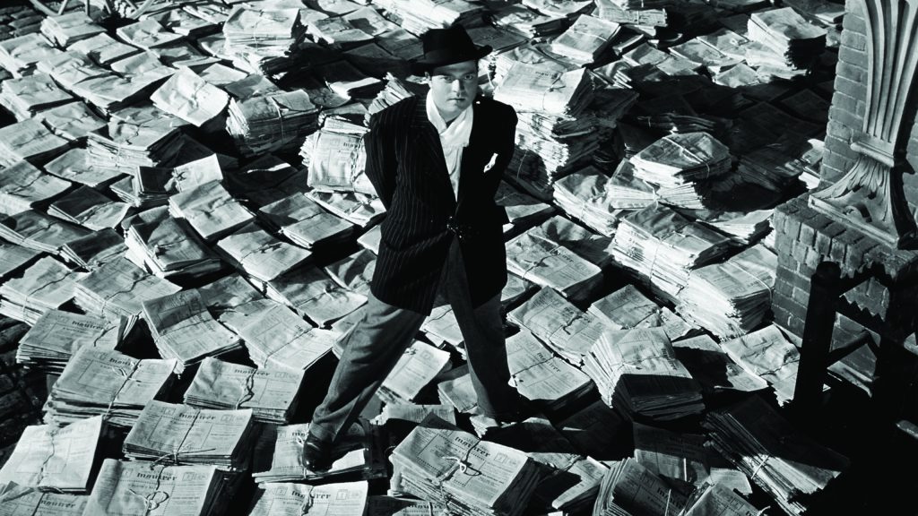 Photo en noir et blanc représentant un homme vêtu d'un costume et d'un borsalino debout sur un tas de journaux.