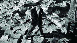 Photo en noir et blanc représentant un homme vêtu d'un costume et d'un borsalino debout sur un tas de journaux.