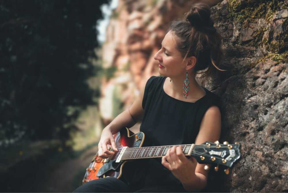 Photo représentant une jeune femme avec un chignon adossée à un arbre en train de jouer de la guitare