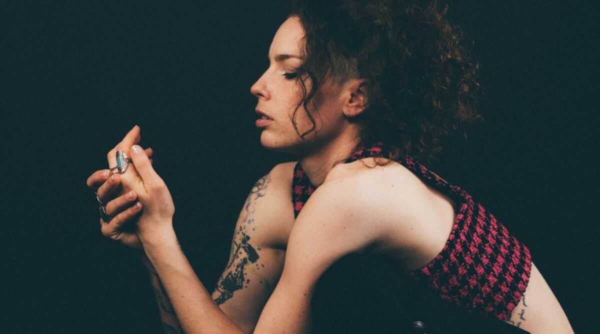Photo représentant une jeune femme de profil en train d'observer une de ses bagues. Elle est tatouée sur le bras et porte une brassière rouge et noir à carreaux.