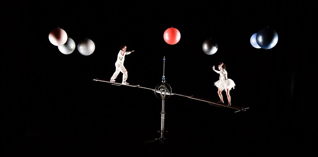 Photo représentant deux circassiens, vêtus de combinaisons de cosmonautes, évoluant en apesanteur autour de planètes, sur une bascule.