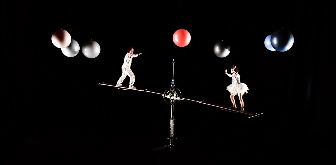 Photo représentant deux circassiens, vêtus de combinaisons de cosmonautes, évoluant en apesanteur autour de planètes, sur une bascule.