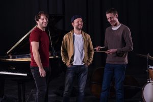 Photo représentant trois musiciens devant un piano en train de sourire.