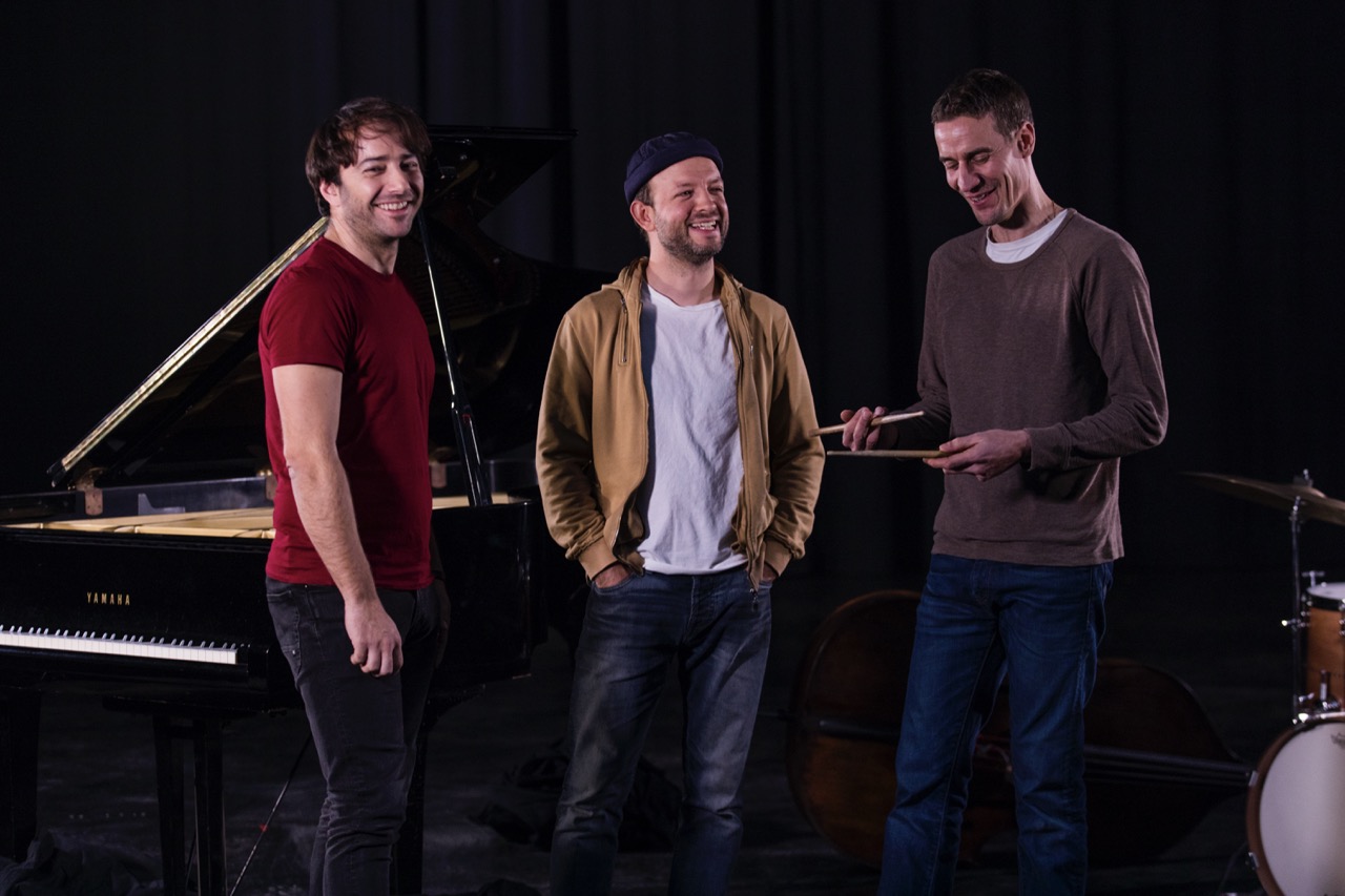 Photo représentant trois musiciens devant un piano en train de sourire.