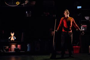Photo représentant sur scène, un comédien vêtu d'un pull rouge regarde vers le haut tandis qu'une marionnette en projection vidéo, ressemblant à un petit renard, apparaît à sa gauche.