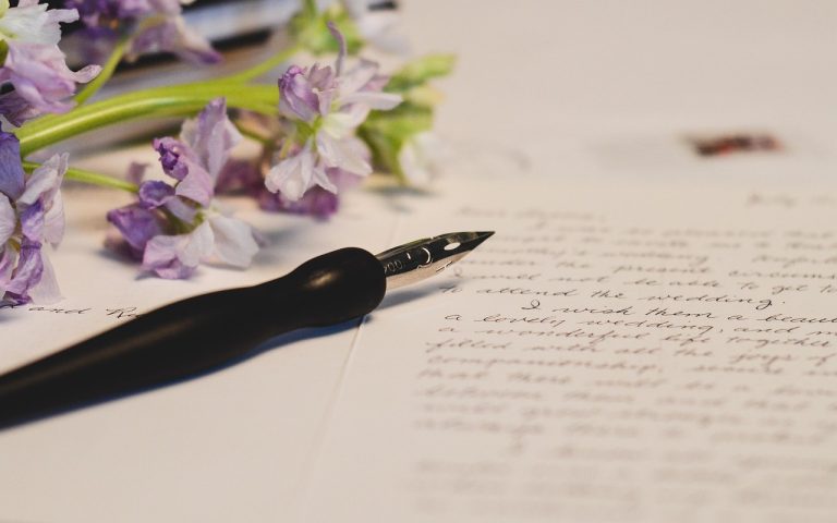 Photo représentant une feuille de papier avec une écriture manuscrite. Dessus on y voit un stylo plume et un bouquet de fleurs violettes